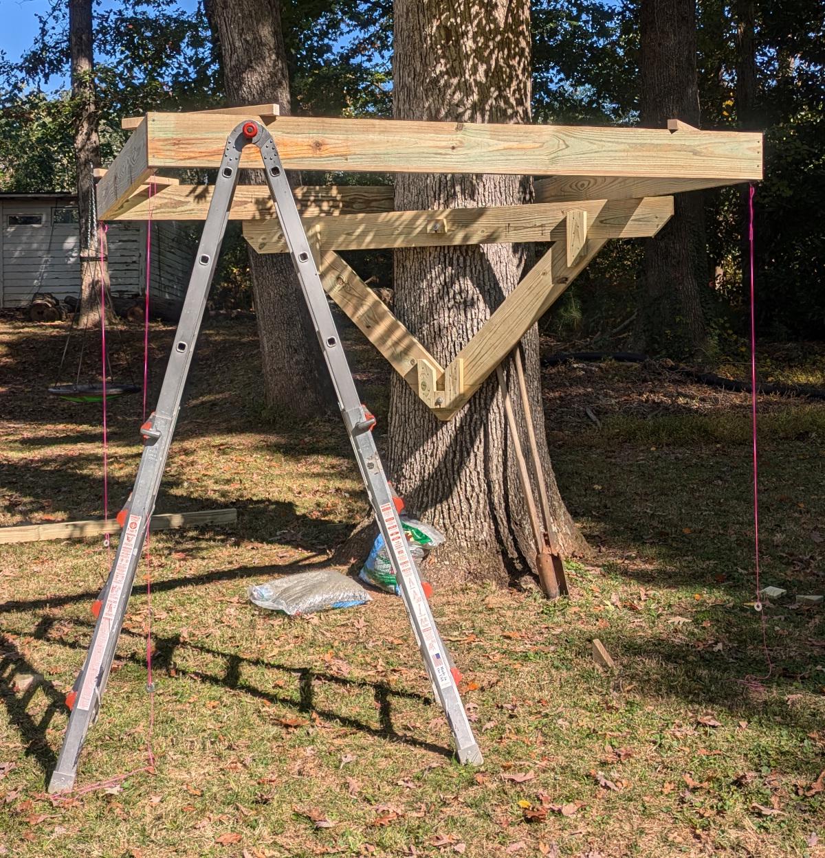 The square joist frame rests on a ladder and the triangular support, which is attached to the tree. The square frame wraps around the tree in one corner and has pink plumb lines extending down from the other corners.