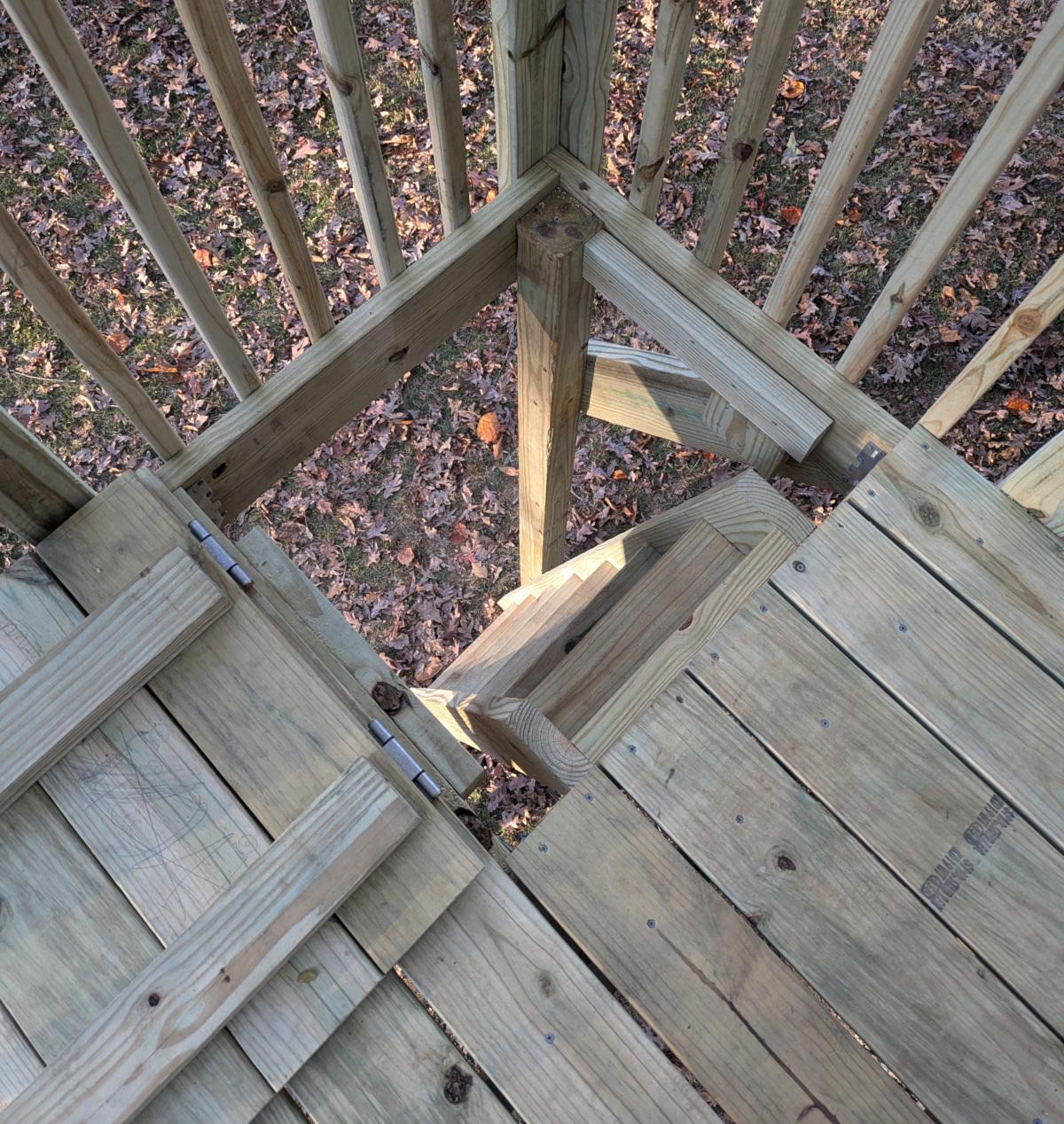 A view of the trap door opening from above. The trap door is attached with two hinges and is visible lying on the decking, open. The ladder is visible through the opening, extending down to the ground at a slight angle.