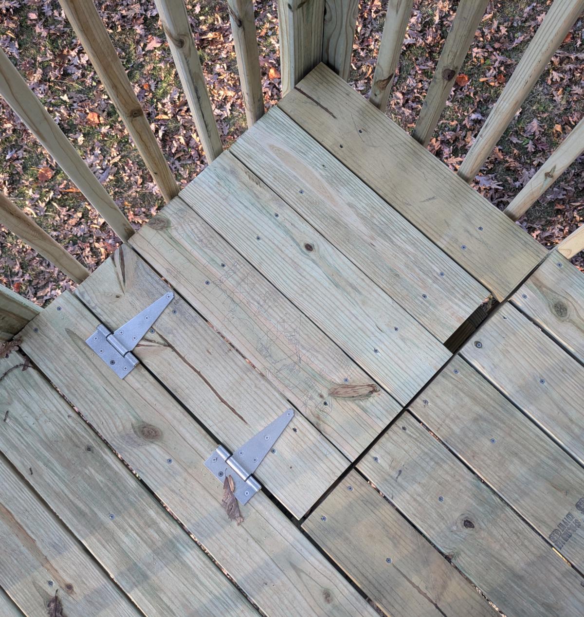 A view of the closed trap door from above. The door is attached with two tee hinges and made of the same decking as the rest of the floor. One piece of decking is slightly shorter than the rest, leaving a gap between it and the floor.