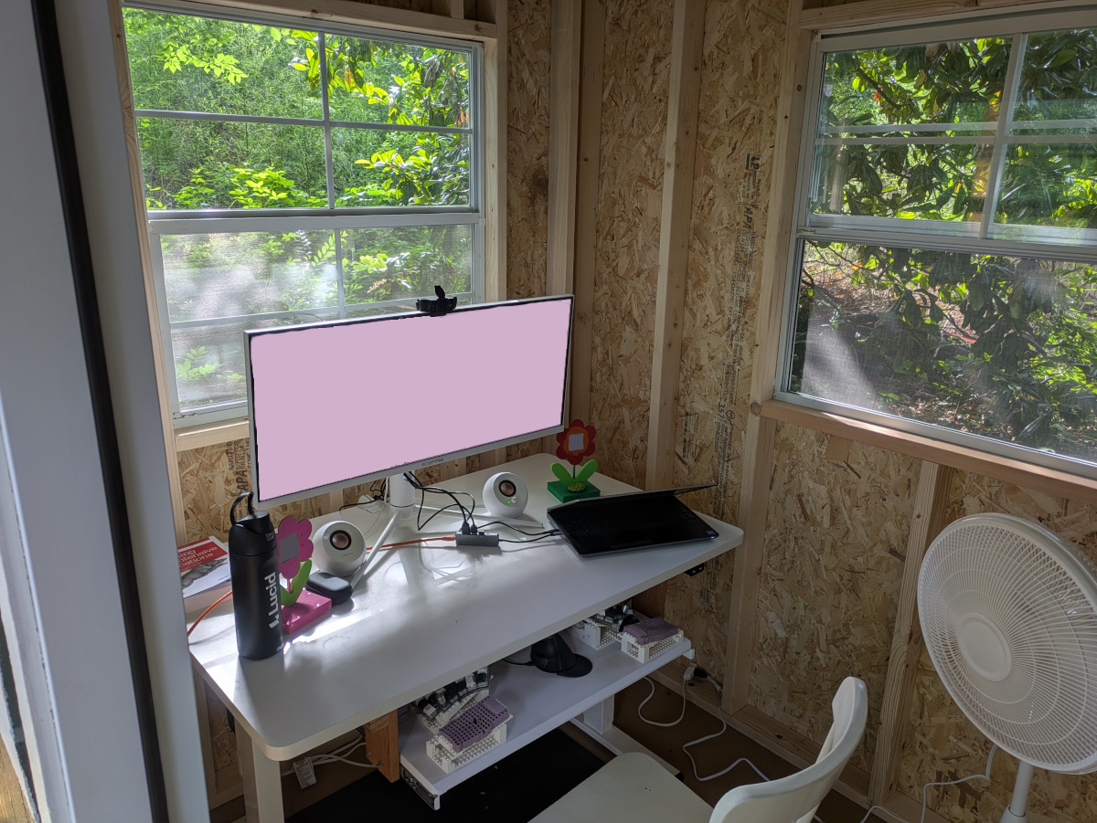 The unfinished interior of the office portion of the shed, with a desk, wide monitor, fan, and several windows.