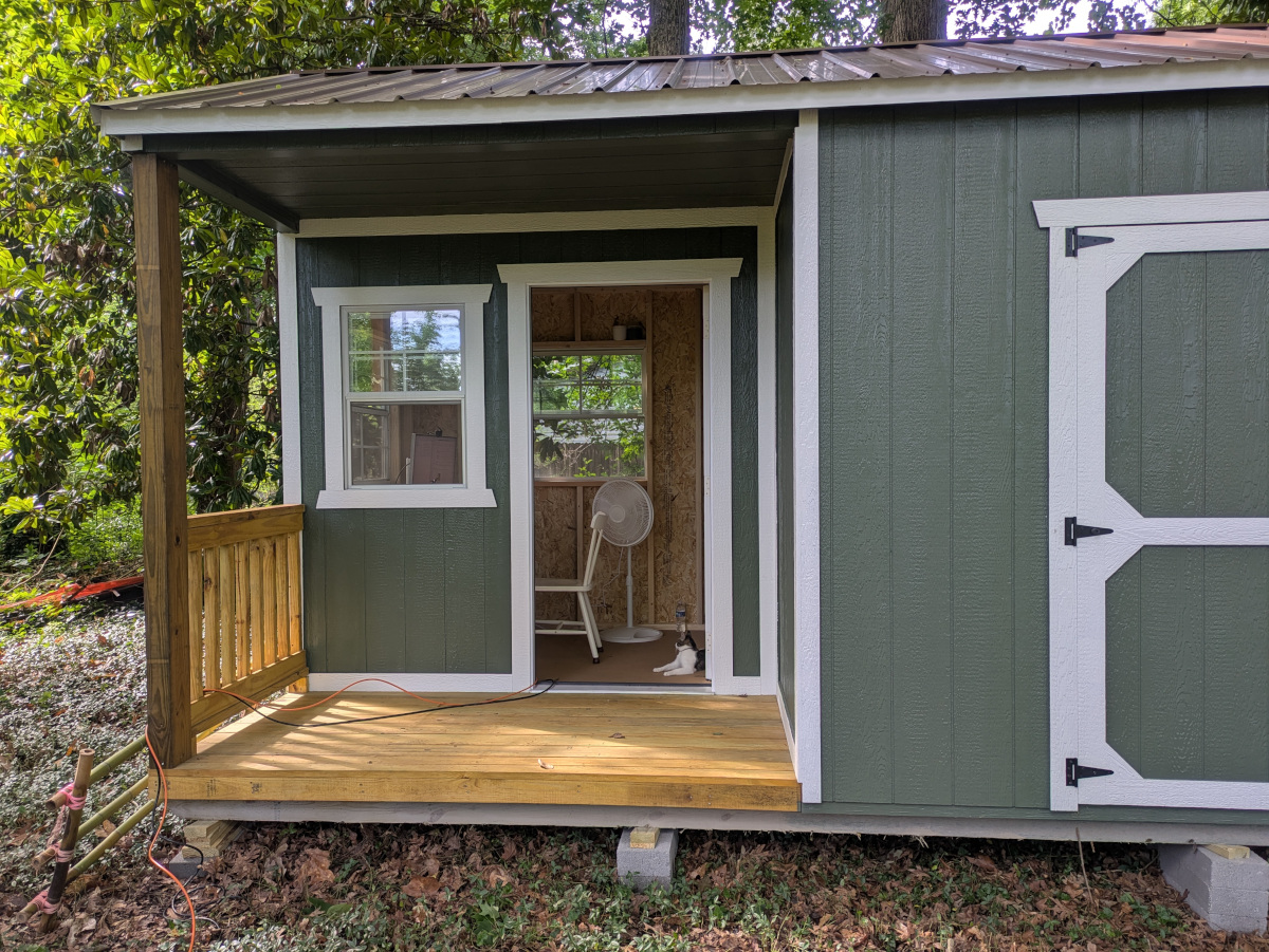 The outside of our shed. A porch is on the left side, with a door open into the office space, and the large door into the storage space is visible on the right.
